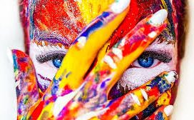 Un niño con la cara y las manos pintadas con muchos colores mira a la cámara tapándose la cara con la mano pintada. 