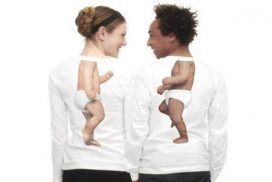 Un hombre y una mujer se miran sonrientes, llevan camisetas que dibujan el cuerpo de dos bebés, niño y niña. 