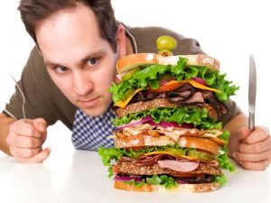 Un hombre mira con ansiedad un sandwich gigante, necesita apoyo psicológico para perder peso y ansiedad.