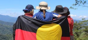 Personas con distintos sombreros y vestidos se abrazan unidos por una bandera. 